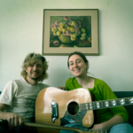 Two people sit smiling on a couch with a guitar and a floral painting in the background
