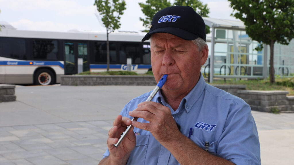 Close-up portrait of Tracy Macdonnell on the right side of the frame, playing a tin whistle with his eyes closed. In the background to the left, a GRT bus is parked at the Cambridge Centre bus station. MacDonnell is wearing a GRT uniform shirt and baseball cap.