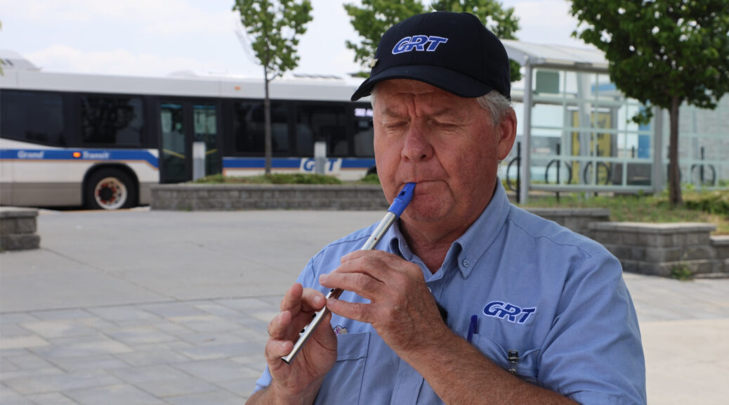 Close-up portrait of Tracy Macdonnell on the right side of the frame, playing a tin whistle with his eyes closed. In the background to the left, a GRT bus is parked at the Cambridge Centre bus station. MacDonnell is wearing a GRT uniform shirt and baseball cap.
