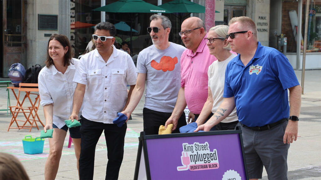 Kitchener City Council members lined up outdoors, getting ready to throw bags at the cornhole competition during the King Street Unplugged opening. From left to right: Stephanie Stretch (Ward 10), Paul Singh (Ward 6), Scott Davey (Ward 1), Mayor Berry Vrbanovic, Debbie Chapman (Ward 9), and Jason Deneault (Ward 3). The photograph shows the council members holding their cornhole bags, set against the background of a downtown stone building.