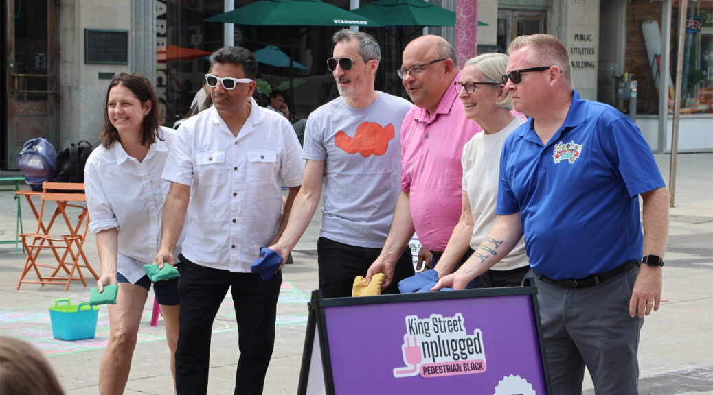 Kitchener City Council members lined up outdoors, getting ready to throw bags at the cornhole competition during the King Street Unplugged opening. From left to right: Stephanie Stretch (Ward 10), Paul Singh (Ward 6), Scott Davey (Ward 1), Mayor Berry Vrbanovic, Debbie Chapman (Ward 9), and Jason Deneault (Ward 3). The photograph shows the council members holding their cornhole bags, set against the background of a downtown stone building.