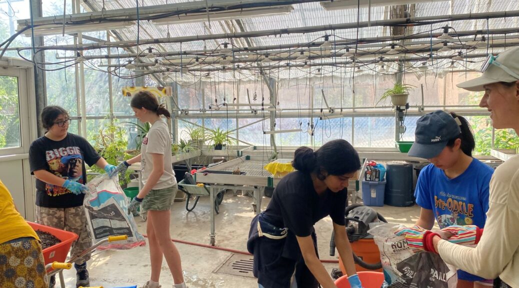5 people are in a greenhouse working with garden m aterials. 4 are youth with one older looking person.