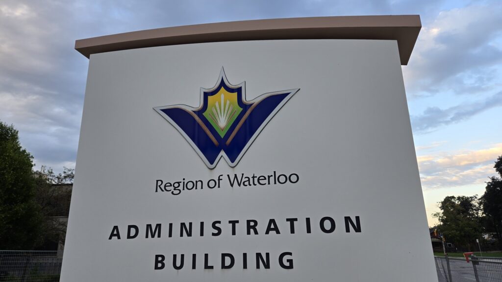 A low-angle outdoor shot of the large, modern white stone monument sign for the Region of Waterloo Administration Building. Centered near the top is the regional logo—a winged, stylized "W" emblem in deep blue, yellow, and green—with the words "Region of Waterloo" directly beneath it, followed by "ADMINISTRATION BUILDING" in bold, black capital letters across two lines, set against green trees and a partly cloudy dusk sky.