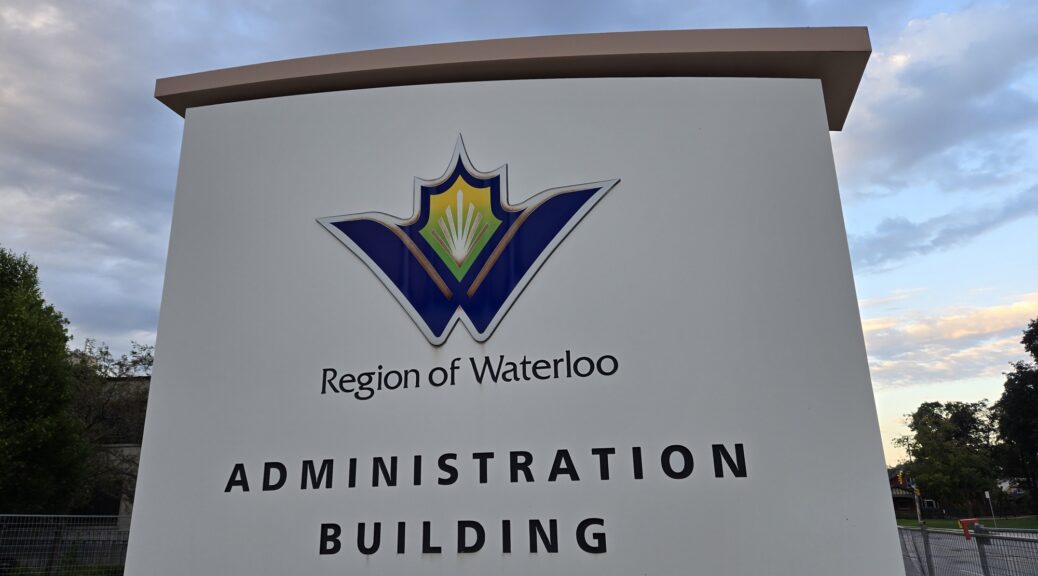 A low-angle outdoor shot of the large, modern white stone monument sign for the Region of Waterloo Administration Building. Centered near the top is the regional logo—a winged, stylized "W" emblem in deep blue, yellow, and green—with the words "Region of Waterloo" directly beneath it, followed by "ADMINISTRATION BUILDING" in bold, black capital letters across two lines, set against green trees and a partly cloudy dusk sky.