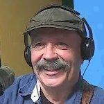 A smiling man with a moustache wearing a flat cap and headphones.