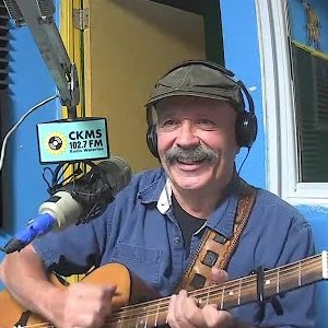 A smiling man with a moustache wearing a flat cap and headphones sits at a microphone and plays guitar.