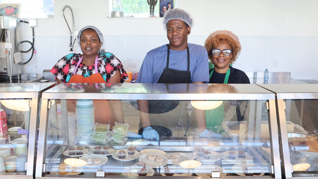 Three volunteers wearing hairnets and aprons stand behind a metal and glass serving counter at St. John's Kitchen in Kitchener, preparing to serve meals to community members in need. From left to right: a woman in a colorful pattern shirt and orange apron, a young man in a blue t-shirt and black apron wearing blue disposable gloves, and a woman with glasses in a green apron. They are smiling at the camera. On display inside the counter glass are plates of cupcakes and donuts, alongside stacked paper cups, with a commercial kitchen set in the background.