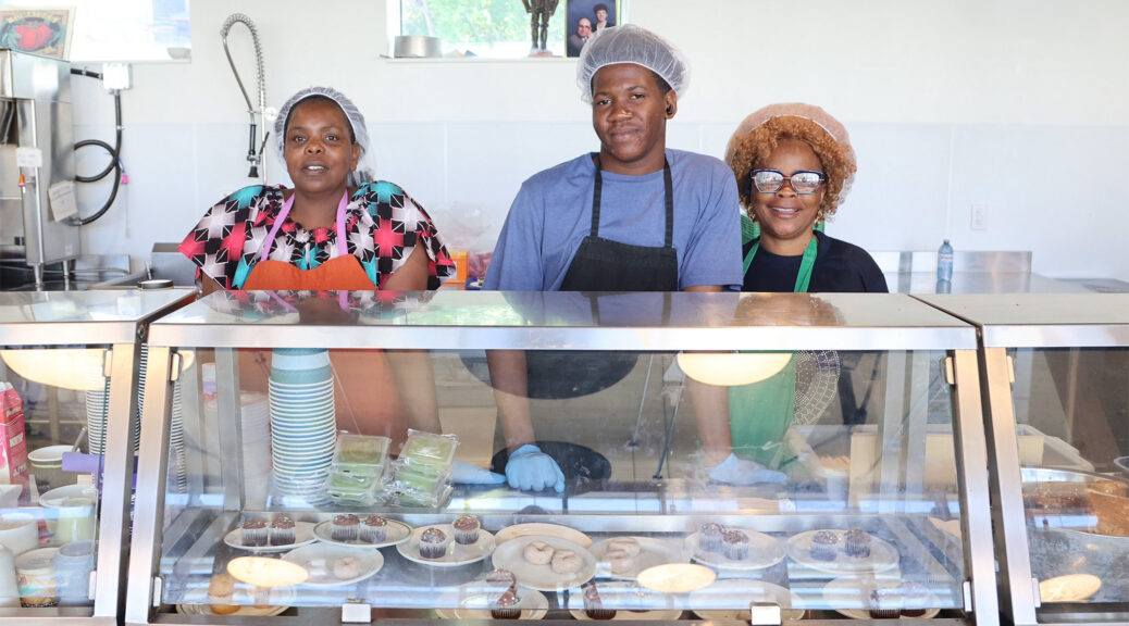 Three volunteers wearing hairnets and aprons stand behind a metal and glass serving counter at St. John's Kitchen in Kitchener, preparing to serve meals to community members in need. From left to right: a woman in a colorful pattern shirt and orange apron, a young man in a blue t-shirt and black apron wearing blue disposable gloves, and a woman with glasses in a green apron. They are smiling at the camera. On display inside the counter glass are plates of cupcakes and donuts, alongside stacked paper cups, with a commercial kitchen set in the background.