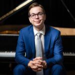 A man in a blue suit sits with a piano behind him and smiles into the camera