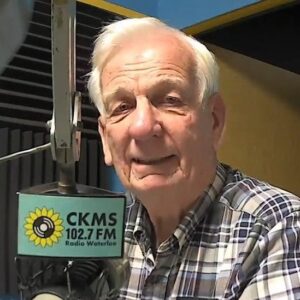 A man with white hair sitting at a microphone looking towards the camera.