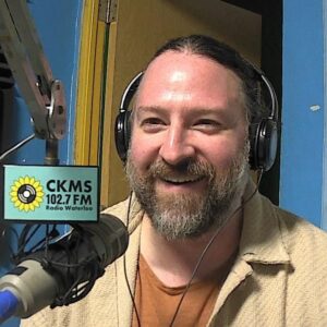 A bearded man wearing headphones sitting at a microphone laughs in the direction of the camera.