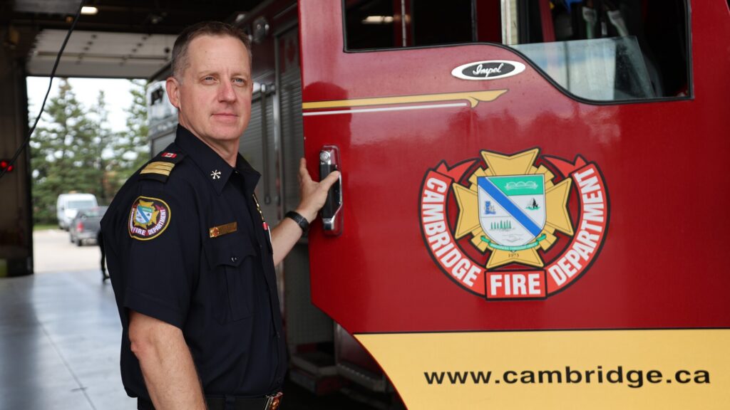 Cambridge Chief Fire Prevention Officer Eric Yates stands in uniform on the left side of the frame inside Station 1, resting his left hand on the open door handle of a red Cambridge Fire Department truck. The large department emblem on the truck door fills the right foreground, while the open bay doors and driveway are visible in the background.