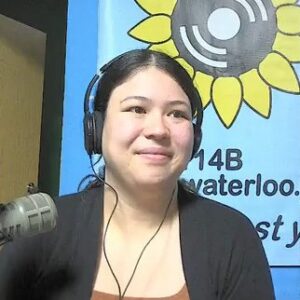 A woman wearing headphones sits at a microphone and smiles to the side of the camera.
