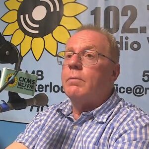 A man with short hair wearing glasses sitting at a microphone with a serious look on his face.