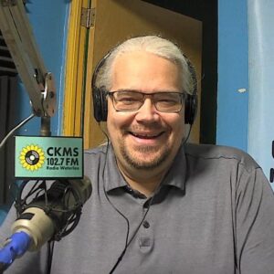 A man wearing headphones, glasses, and a gray shirt sitting next to a microphone smiles towards the camera.