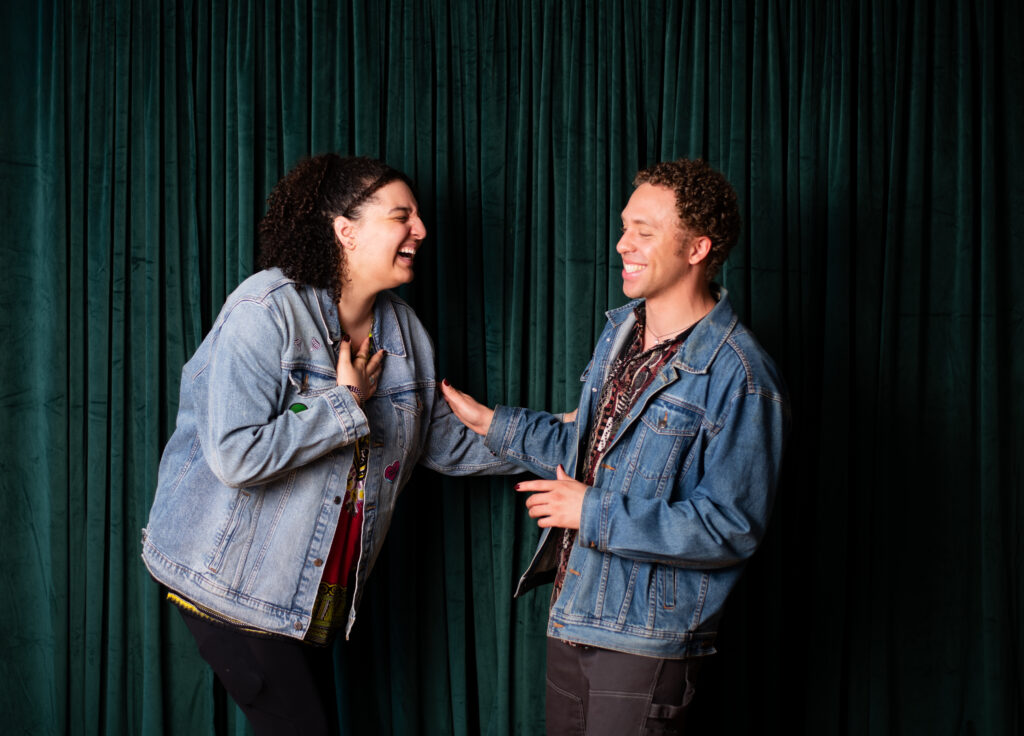 Two biracial people wearing jean jackets laugh together in front of a green velvet curtain.