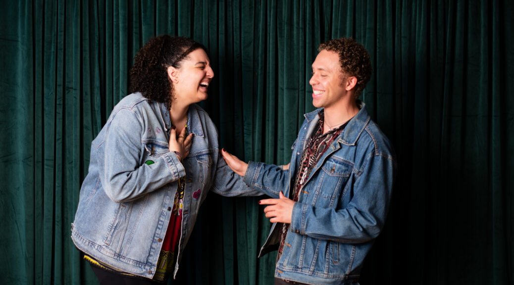 Two biracial people wearing jean jackets laugh together in front of a green velvet curtain.