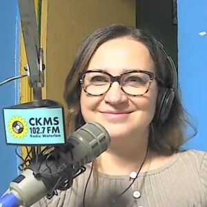 A woman wearing headphone in front of a microphone smiling towards the camera.