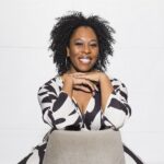 A woman wearing a black and white dress sits backwards on a chair and smiles into the camera