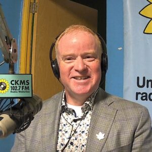 A man wearing headphones and a checkered jacket sitting at a microphone smiles towards the camera.