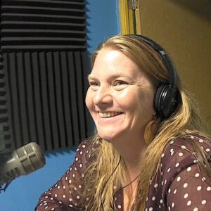A woman with long blonde hair wearing headphones sits at a microphone and smiles towards the left.