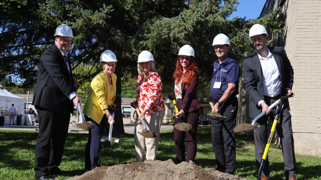 Six officials wearing white hard hats stand in a row on a lawn during a groundbreaking ceremony, holding shovels over a pile of freshly dug earth. The participants include individuals in formal suits, a yellow blazer, a floral top, and a plum pantsuit. The background features tall evergreen trees and the exterior of a brick building.