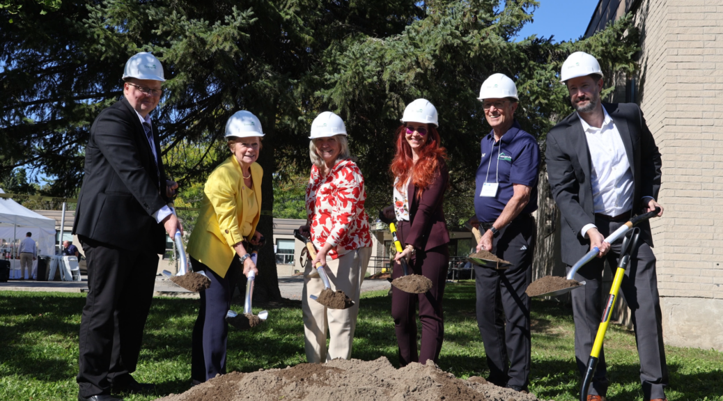 Six officials wearing white hard hats stand in a row on a lawn during a groundbreaking ceremony, holding shovels over a pile of freshly dug earth. The participants include individuals in formal suits, a yellow blazer, a floral top, and a plum pantsuit. The background features tall evergreen trees and the exterior of a brick building.