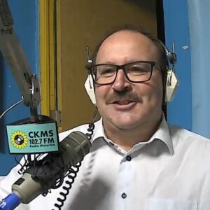 A man with a moustache wearing glasses and headphones sitting at a microphone and smiling towards the camera.