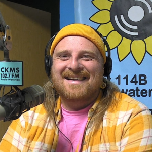 A bearded man wearing a pink T-shirt, yellow checkered lumberjack shirt, a yellow toque, and headphones sits at a microphone and smiles towards the camera.