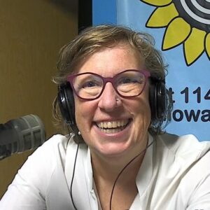 A woman with short curly hair wearing glasses and headphones laughs towards the camera.