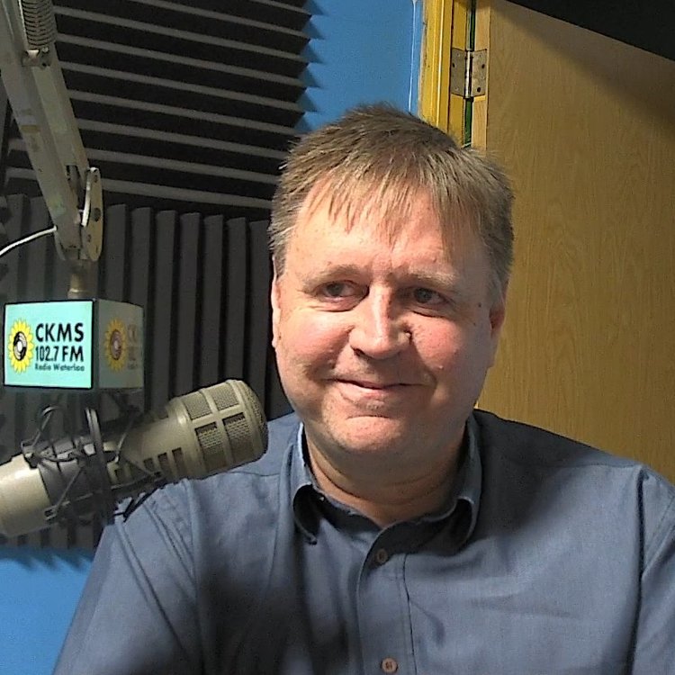 A man wearing a blue shirt sitting at a microphone smiles towards the camera