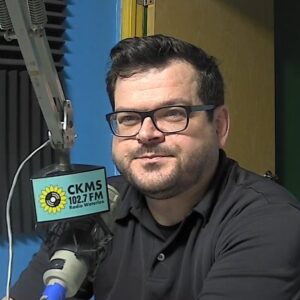 A man with a beard and moustache wearing glasses, sitting at a microphone looks towards the camera.