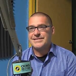 A man with very short hair wearing glasses sits at a microphone and smiles towards the camera.