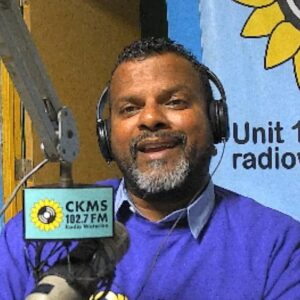 A bearded man wearing headphones sitting at a microphone speaking towards the camera