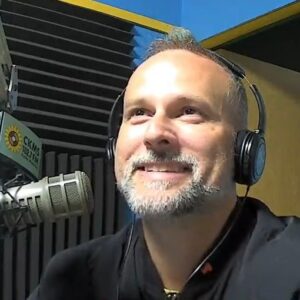 A man with a goatee and wearing headphones sitting at a microphone smiles upwards.