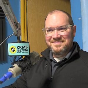 A man with beard and moustache, wearing glasses and a brown jacket sits in front of a microphone and smiles towards the camera