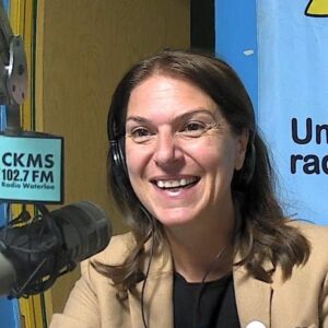 A woman with long hair wearing a taupe jacket sits at a microphone and smiles towards the camera
