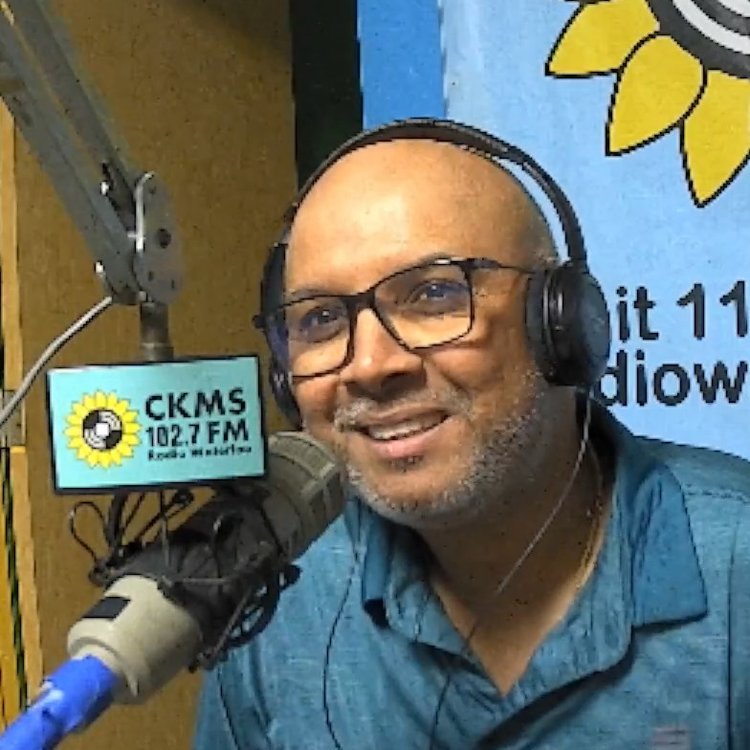 A bald man with beard stubble wearing a blue shirt and headphones leaning towards a microphone and smiling towards the camera.
