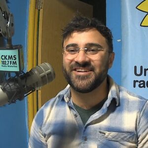 A bearded man wearing a blue plaid shirt and glasses sits at a microphone and smiles towards the camera.