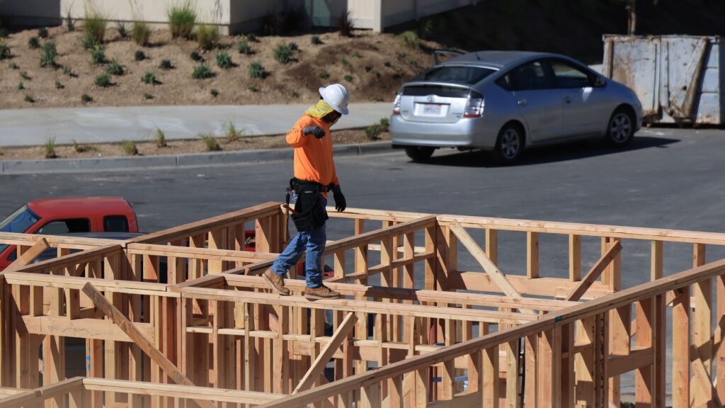 A high-angle photograph of a construction worker wearing a white hard hat, a yellow face cover, an orange long-sleeve shirt, blue jeans, and a tool belt, walking carefully on top of the open wooden wall frames of a multi-room residential building under construction. In the background, a residential area is visible with an open-door silver hatchback parked on an asphalt driveway, alongside a red vehicle partially visible in the lower left, scattered shrubs, and a metal dumpster near the top right. The daylight is bright, casting shadows of the wood framing on the ground.