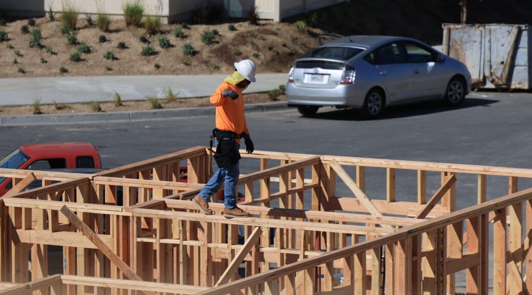 A high-angle photograph of a construction worker wearing a white hard hat, a yellow face cover, an orange long-sleeve shirt, blue jeans, and a tool belt, walking carefully on top of the open wooden wall frames of a multi-room residential building under construction. In the background, a residential area is visible with an open-door silver hatchback parked on an asphalt driveway, alongside a red vehicle partially visible in the lower left, scattered shrubs, and a metal dumpster near the top right. The daylight is bright, casting shadows of the wood framing on the ground.